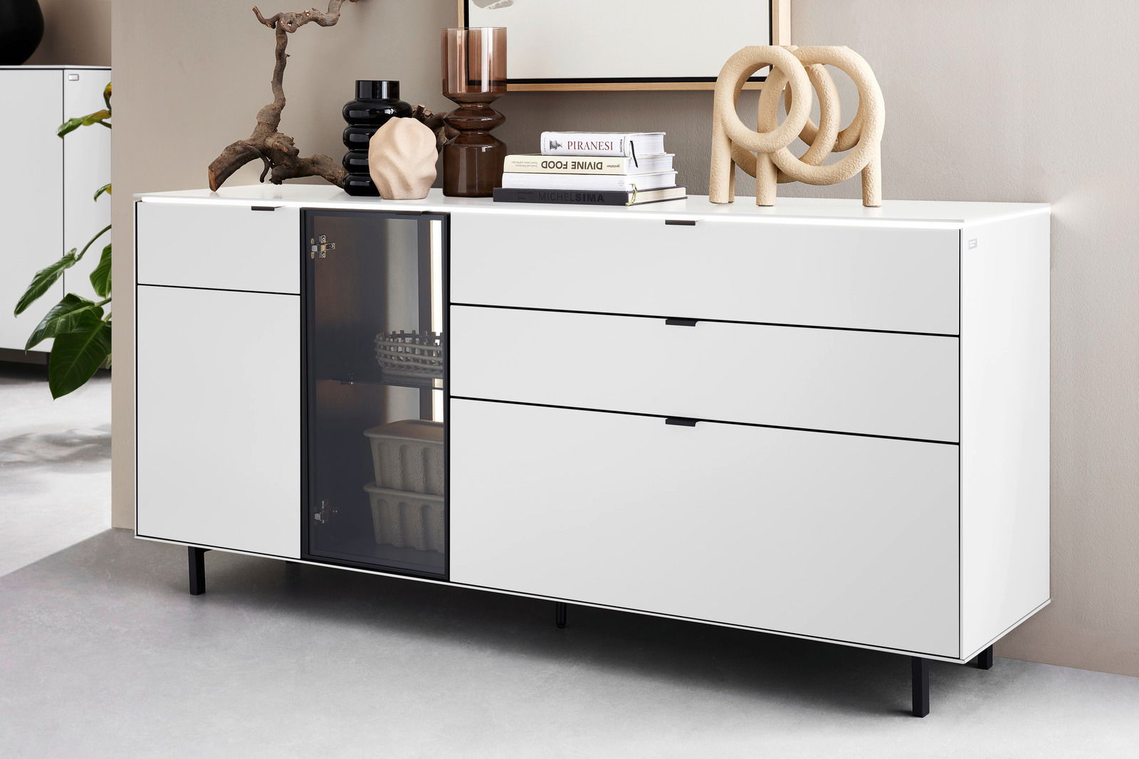 Modernes weißes Sideboard mit drei Schubladen und einer Glastür, dekoriert mit Büchern und Skulpturen, seitliche Perspektive.