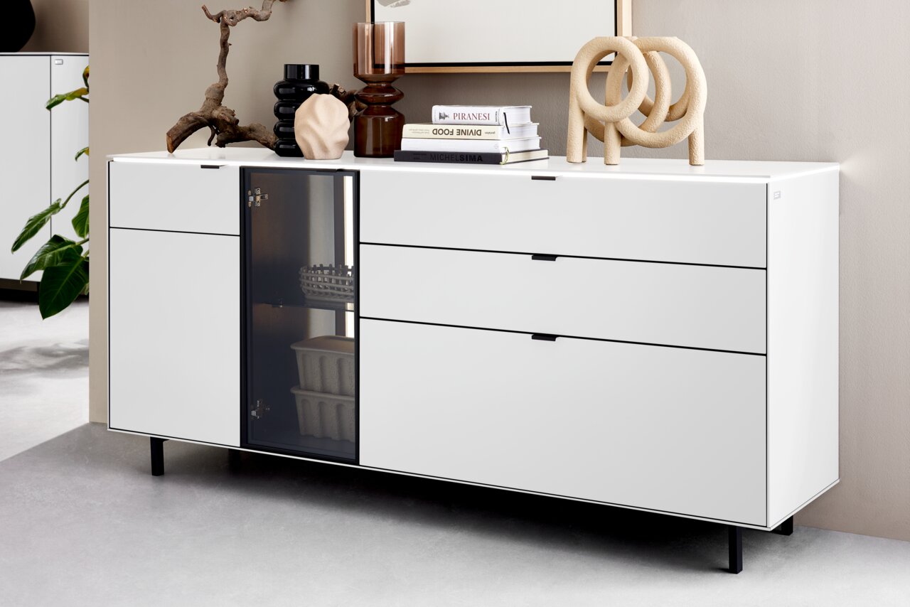 Modernes weißes Sideboard mit drei Schubladen und einer Glastür, dekoriert mit Büchern und Skulpturen, seitliche Perspektive.