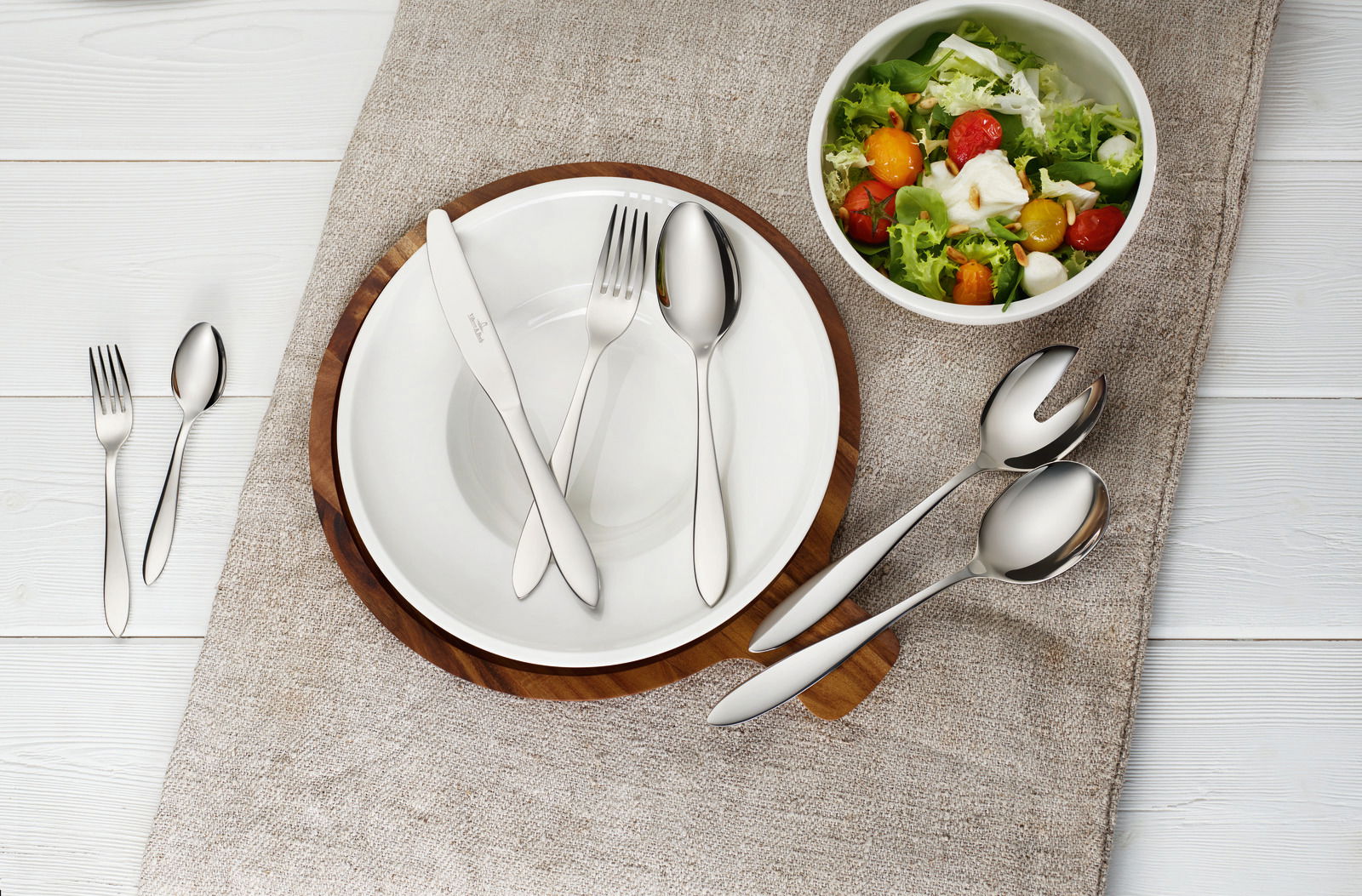 Vogelperspektive auf ein gedecktes Tischset mit Besteck Arthur 68-tlg., bestehend aus Messer, Gabel und Löffel auf einem weißen Teller, daneben eine Schüssel mit Salat.