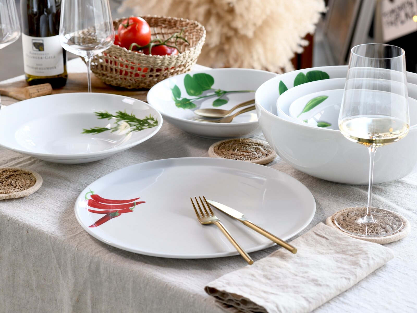 Schlichtes weißes Geschirrset mit botanischen Motiven (Chili, Kräuter, grüne Blätter) auf gedecktem Tisch mit Weingläsern und goldfarbenem Besteck, fotografiert aus leicht erhöhter schräger Perspektive.