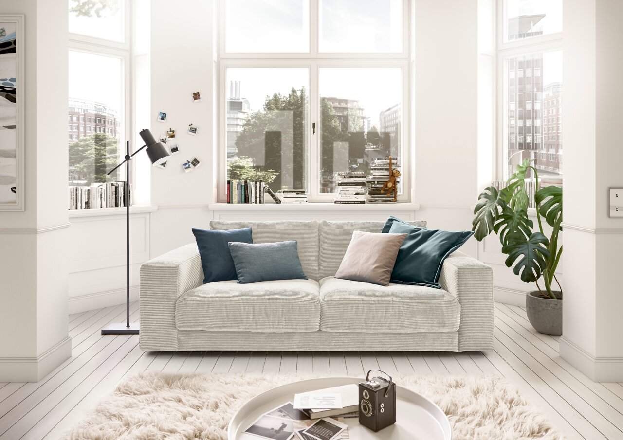 Zweisitzer-Sofa in einem hellen Wohnzimmer mit großen Fenstern, dekoriert mit Kissen in Blau- und Grautönen, aus der Frontalperspektive.
