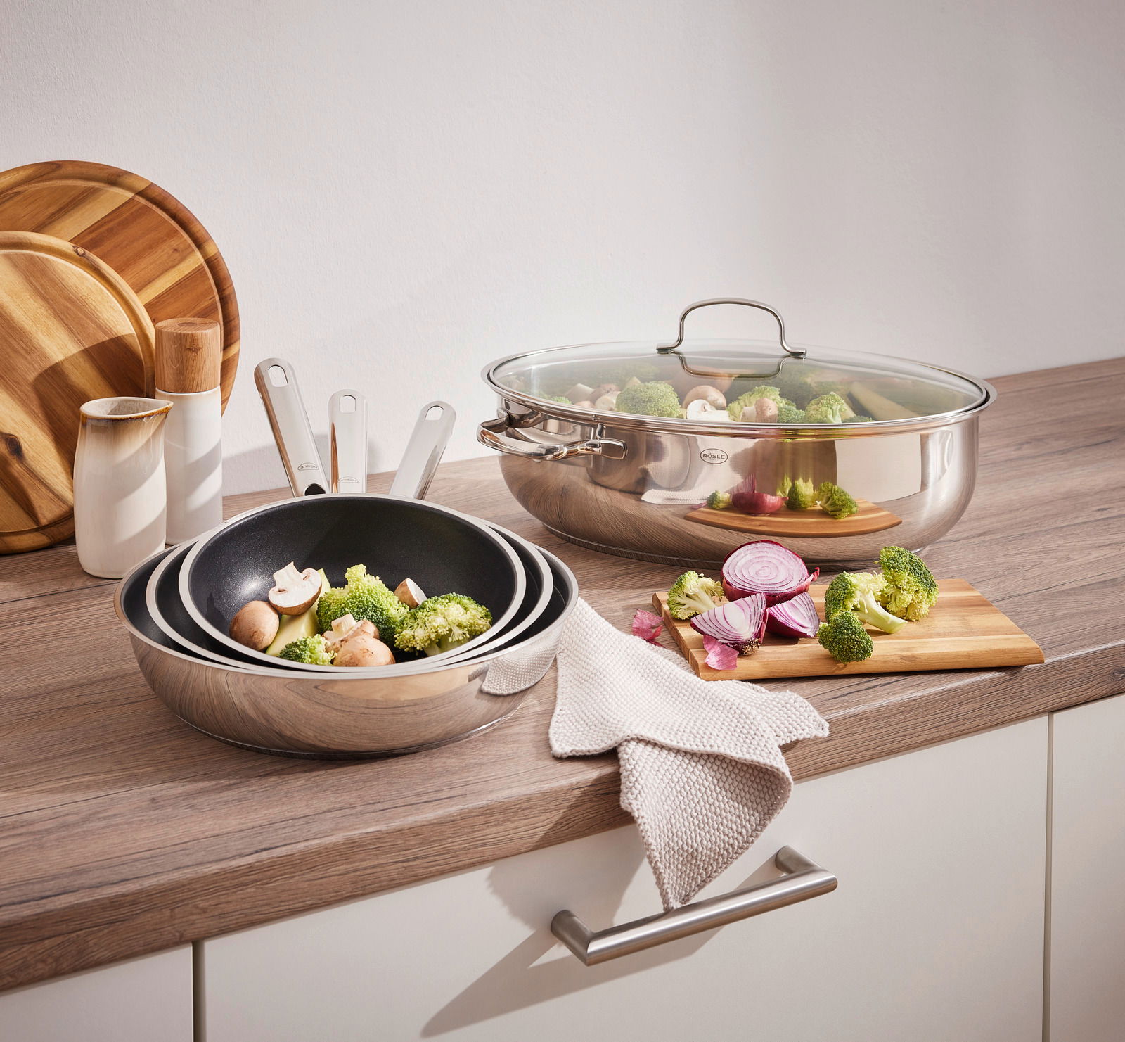 Rösle Pfannenset 3-tlg. ELEGANCE Pfannenset Elegance 3-tlg. auf einer Küchenarbeitsplatte, bestehend aus zwei Pfannen und einem Bräter mit Glasdeckel, gefüllt mit frischem Gemüse. Perspektive von vorne.