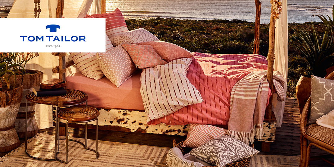 Bed met roze beddengoed en veel kussens in de vrije natuur aan zee. Logo Tom Tailor