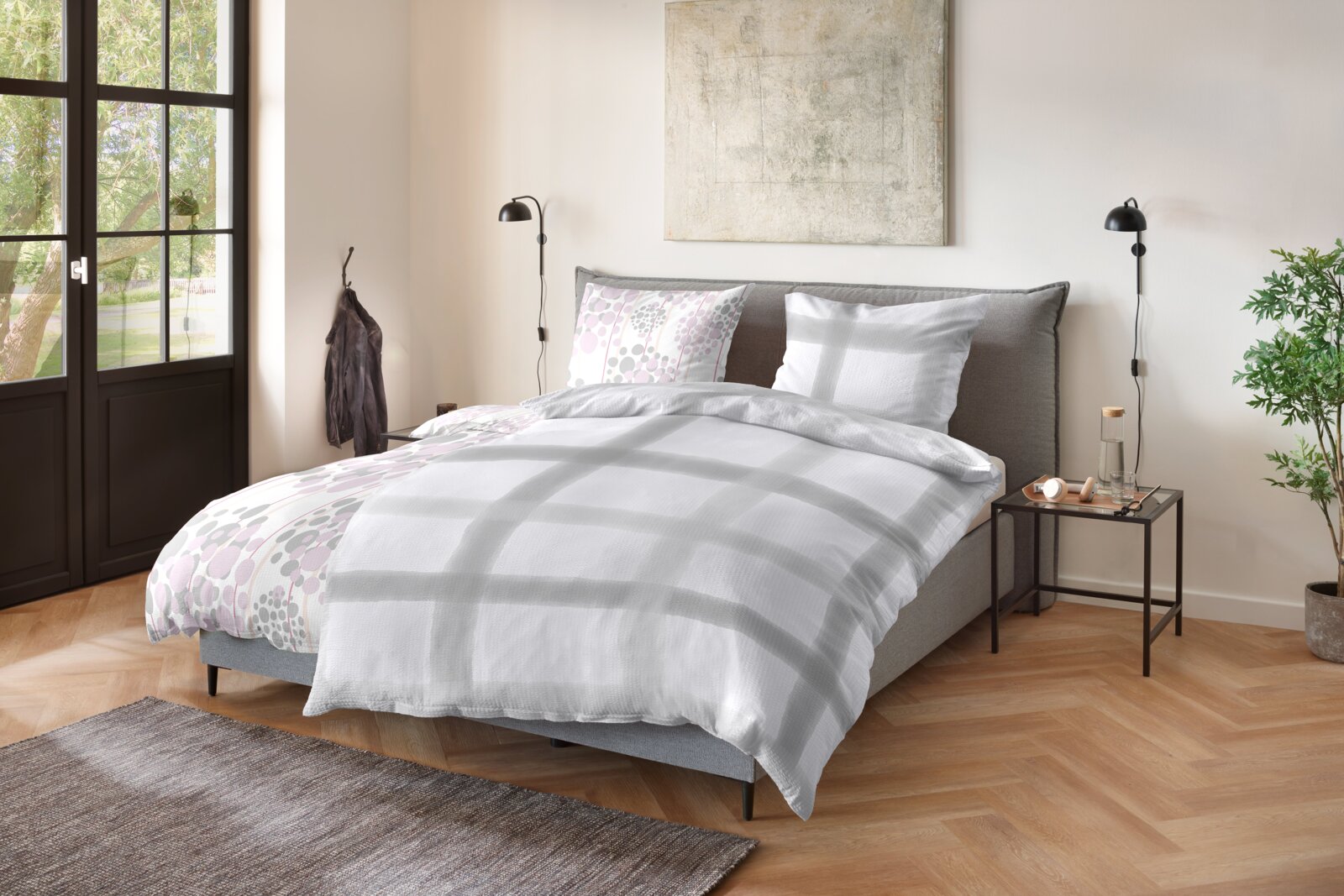 Primera Bettwäsche Modernes Schlafzimmer mit grauem Polsterbett und Bettwäsche in Weiß mit grauen Karos sowie pastellfarbenem Punkte-Muster; Ansicht schräg von vorne aus leicht erhöhter Perspektive.