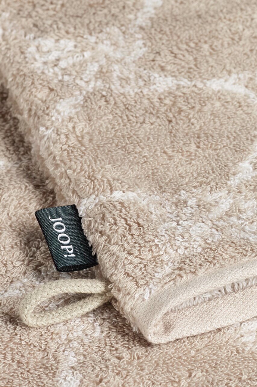JOOP! Waschhandschuh CORNFLOWER CLASSIC Nahaufnahme eines beigen Waschhandschuhs mit einem schwarzen Etikett mit der Aufschrift 'JOOP!' und einer Schlaufe, liegend auf einer flauschigen Oberfläche.