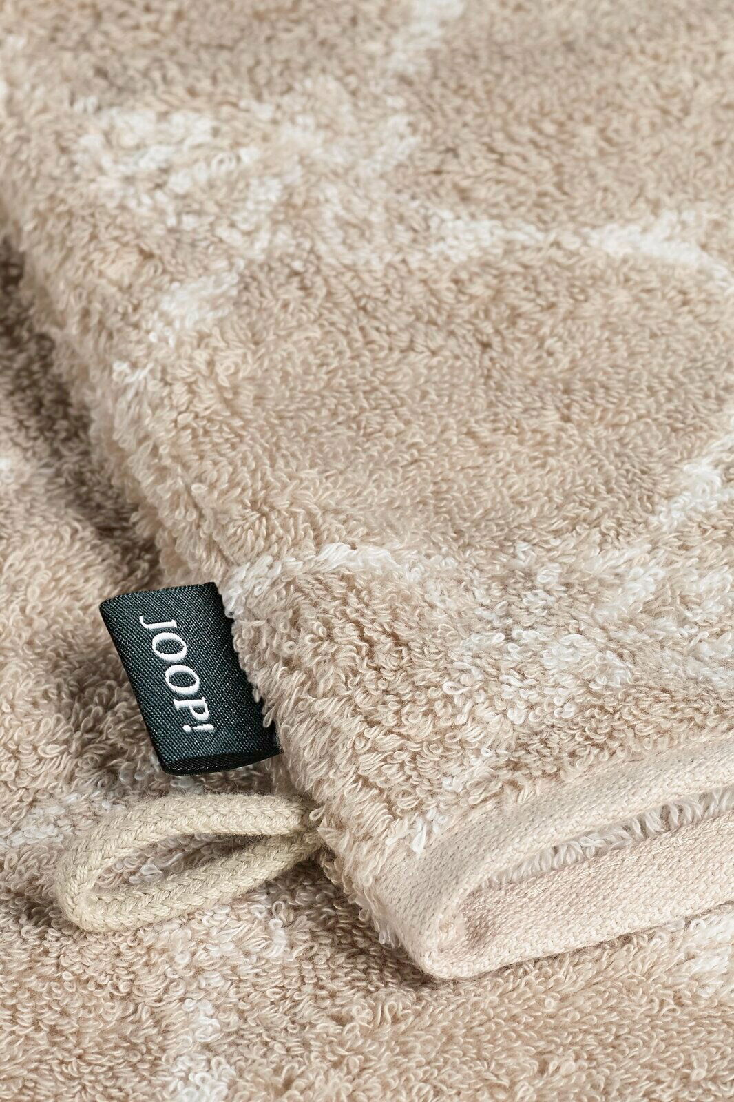 JOOP! Waschhandschuh CORNFLOWER CLASSIC Nahaufnahme eines beigen Waschhandschuhs mit einem schwarzen Etikett mit der Aufschrift 'JOOP!' und einer Schlaufe, liegend auf einer flauschigen Oberfläche.