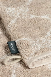 Nahaufnahme eines beigen Waschhandschuhs mit einem schwarzen Etikett mit der Aufschrift 'JOOP!' und einer Schlaufe, liegend auf einer flauschigen Oberfläche.