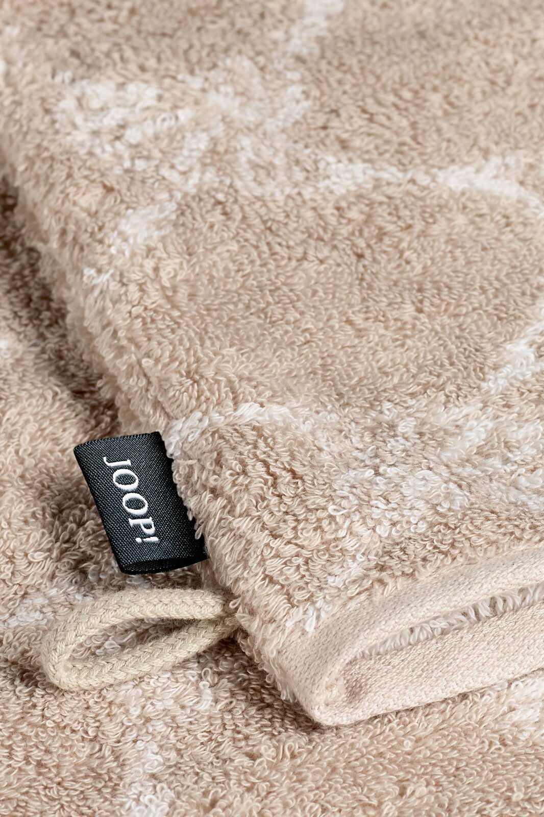 JOOP! Waschhandschuh CORNFLOWER CLASSIC Nahaufnahme eines beigen Waschhandschuhs mit einem schwarzen Etikett mit der Aufschrift 'JOOP!' und einer Schlaufe, liegend auf einer flauschigen Oberfläche.