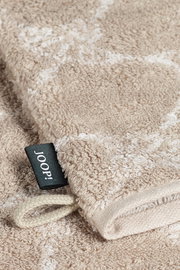 Nahaufnahme eines beigen Waschhandschuhs mit einem schwarzen Etikett mit der Aufschrift 'JOOP!' und einer Schlaufe, liegend auf einer flauschigen Oberfläche.