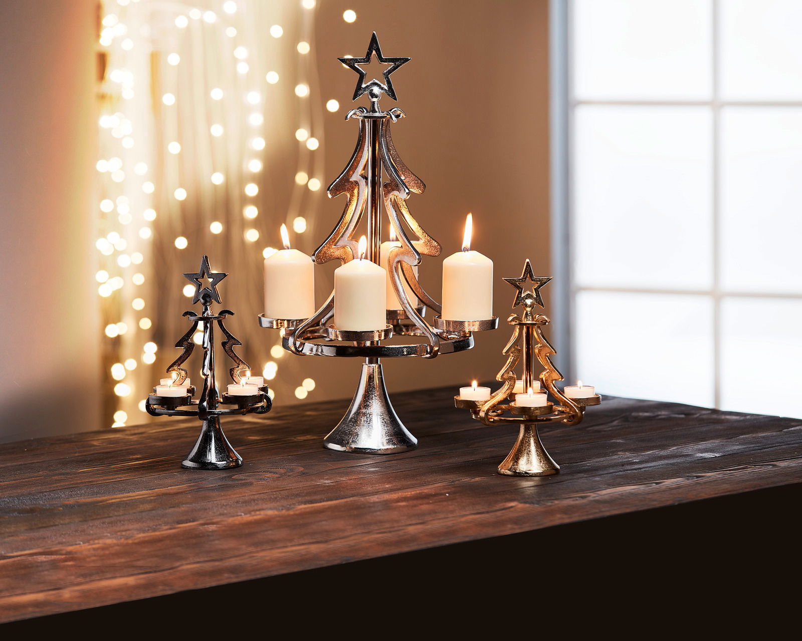 Kerzenhalter in Form eines Tannenbaums aus Aluminium, in verschiedenen Größen und Farben, mit brennenden Kerzen, auf einem Tisch vor einem Fenster mit Lichterkette im Hintergrund, seitliche Perspektive.