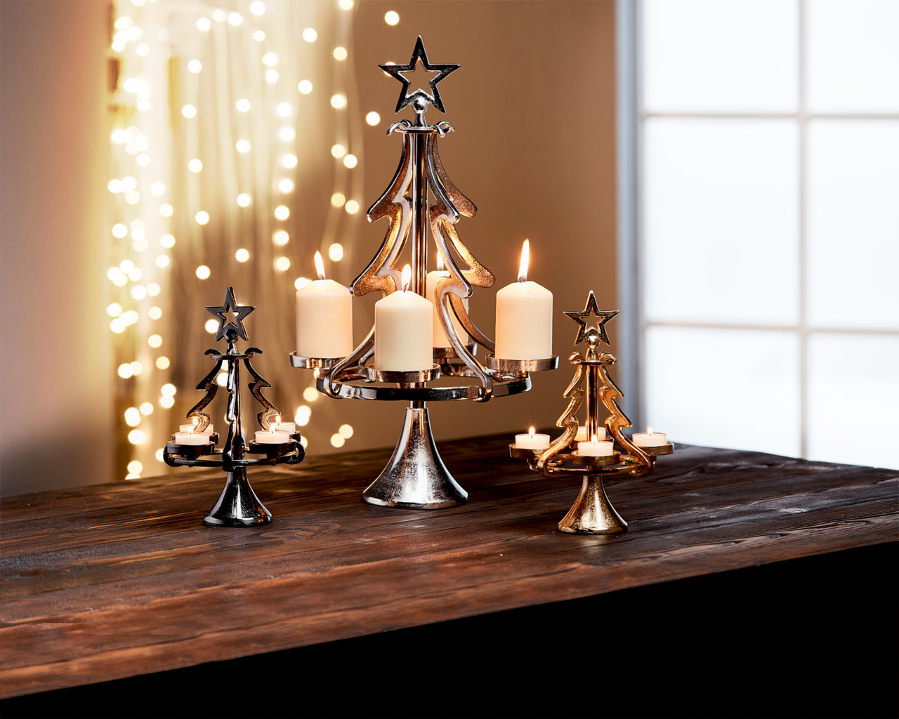 Kerzenhalter in Form eines Tannenbaums aus Aluminium, in verschiedenen Größen und Farben, mit brennenden Kerzen, auf einem Tisch vor einem Fenster mit Lichterkette im Hintergrund, seitliche Perspektive.