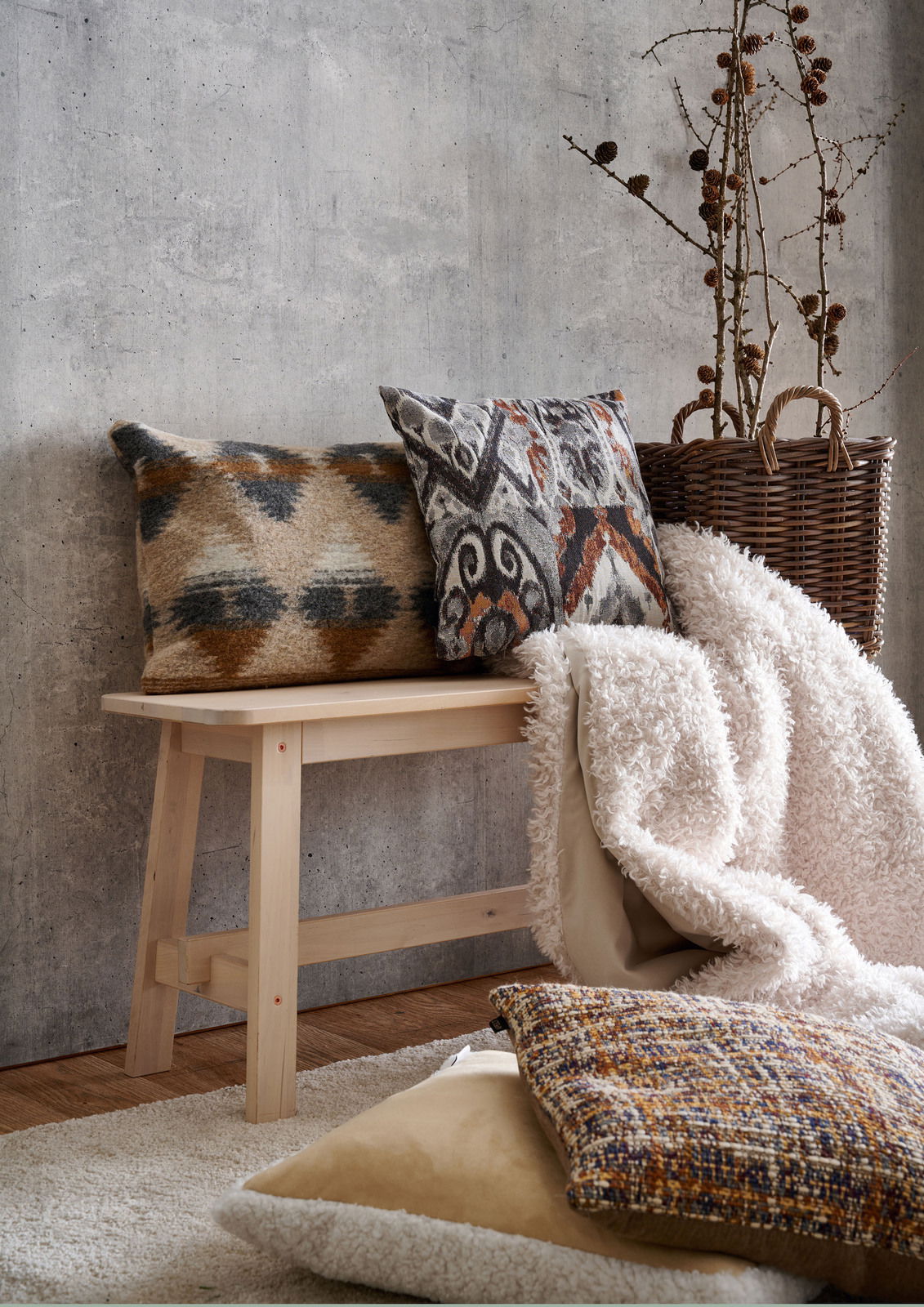Apelt Kissenhülle SIERRA Gemütliche Wohnszene mit zwei dekorativen Kissen auf einer Holzbank, umgeben von einem Korb mit Zweigen und einer flauschigen Decke, aus seitlicher Perspektive fotografiert.