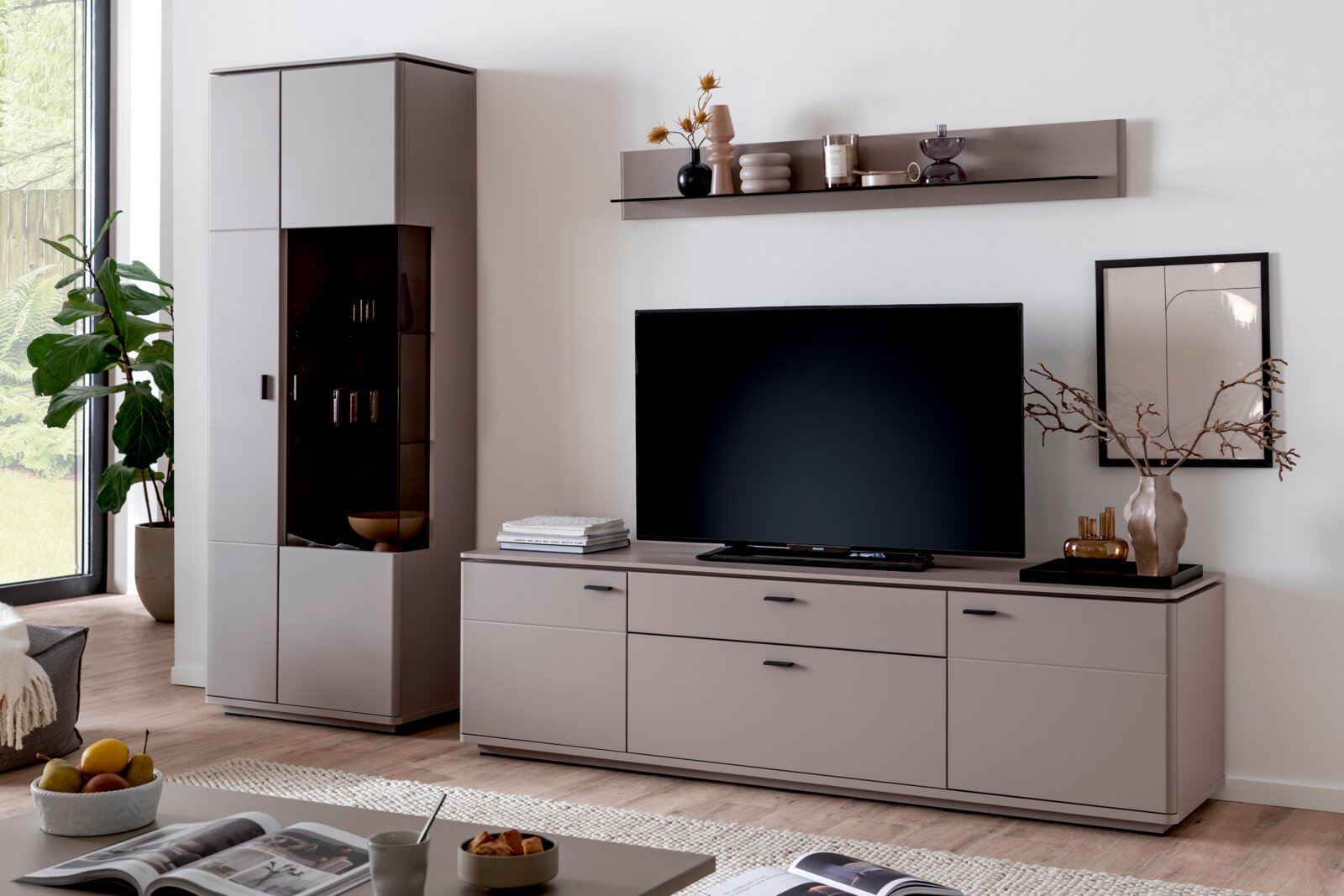 Zurbrüggen Wandkombination TORRENT Modernes Wohnzimmer-Setup mit grauem TV-Lowboard und passendem Hochschrank mit Glasvitrine, TV mittig platziert und Wandboard darüber; Frontansicht in leichter Schrägperspektive von links.