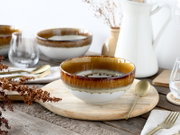 Keramikschale in Braun-Weiß mit reaktiver Glasur auf rundem Holzbrett, mit goldenem Löffel auf rustikalem Holztisch, frontal aus leicht erhöhter Perspektive fotografiert.