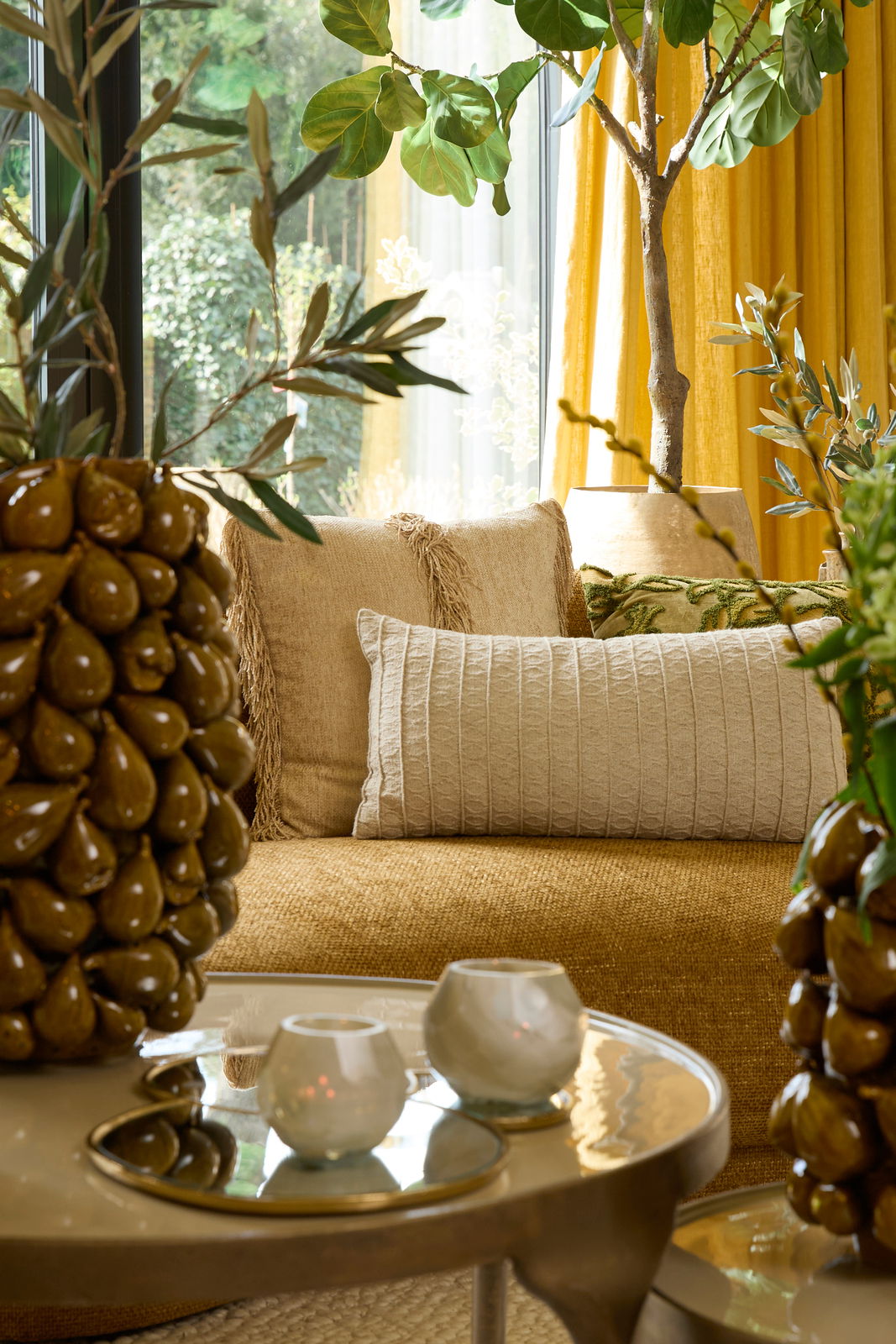 Ein gemütliches Wohnzimmer mit einem beigen Kissen auf einem Sofa, umgeben von Pflanzen und einem Tisch mit Kerzenhaltern. Perspektive: seitlich.