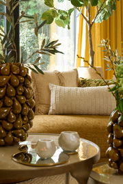 Ein gemütliches Wohnzimmer mit einem beigen Kissen auf einem Sofa, umgeben von Pflanzen und einem Tisch mit Kerzenhaltern. Perspektive: seitlich.