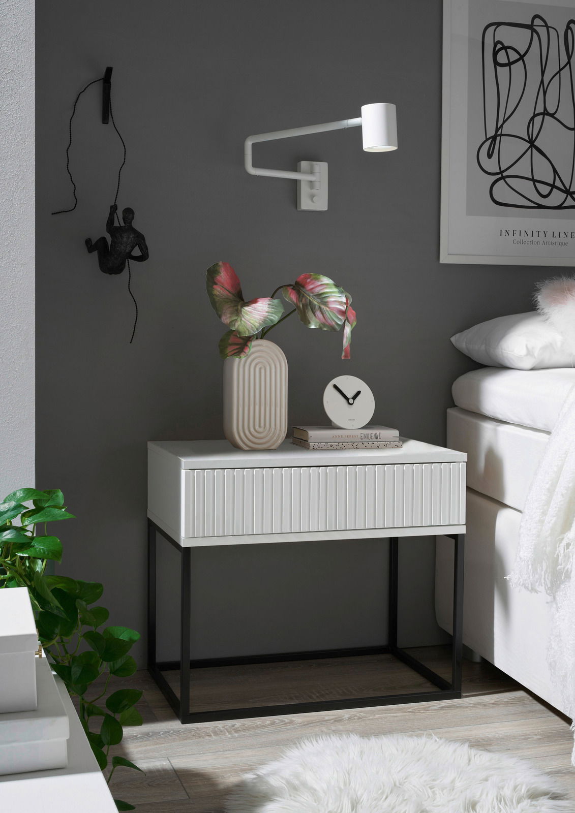 Moderne Nachtkommode mit weißer Oberfläche und schwarzen Metallbeinen, dekoriert mit einer Vase und einer Uhr, seitliche Perspektive.