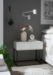 Moderne Nachtkommode mit weißer Oberfläche und schwarzen Metallbeinen, dekoriert mit einer Vase und einer Uhr, seitliche Perspektive.