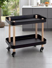 Schwarzer, zweistöckiger Servierwagen mit zwei Tablettablagen, hellen Holzpfosten und vier Rollen in moderner Küche, schräg von vorn fotografiert.