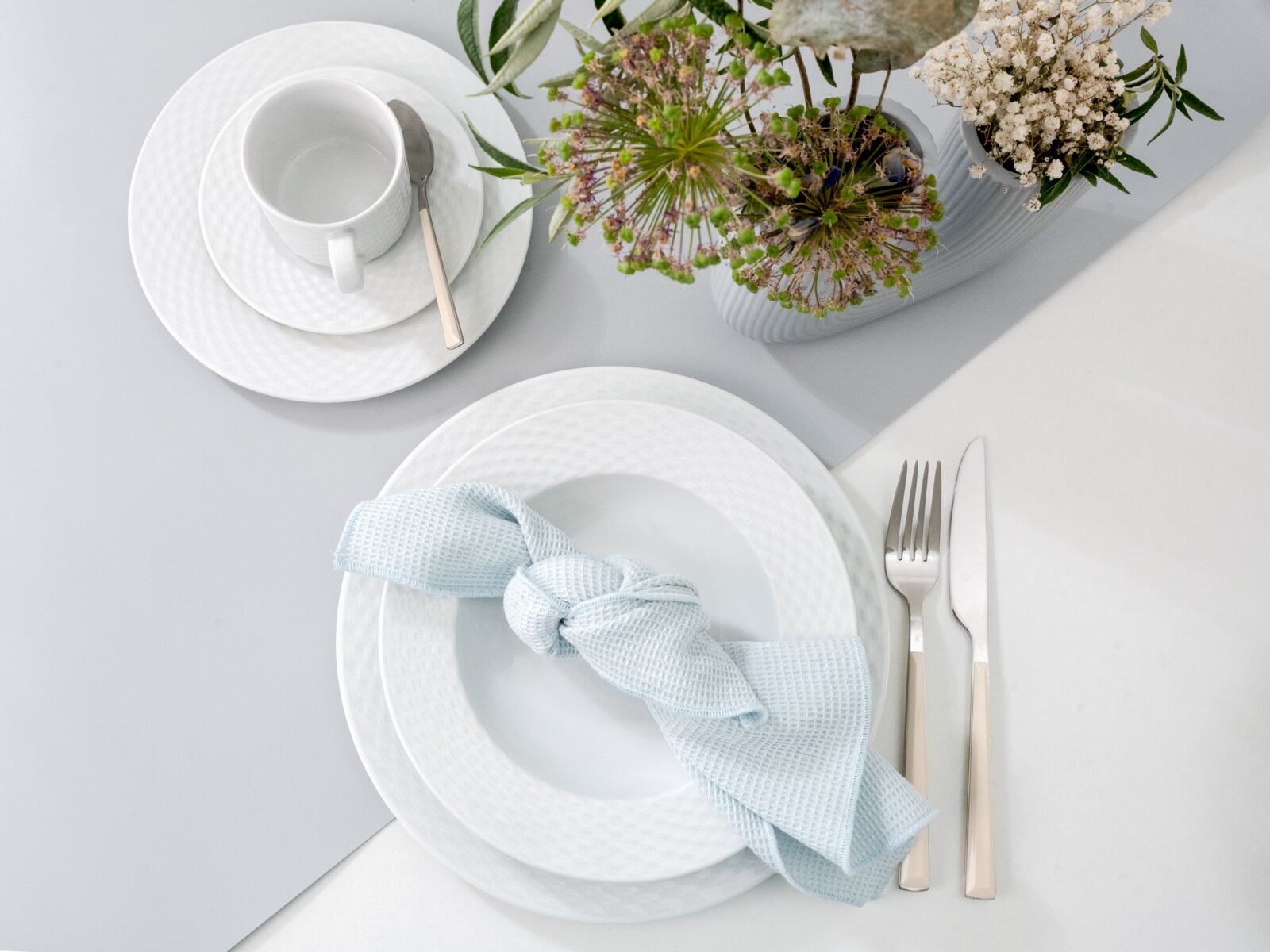 Minimalistisches weißes Geschirr-Arrangement: Tasse auf Untertasse mit Löffel, tiefer Teller mit hellblauer Stoffserviette sowie Messer und Gabel neben einer weißen Vase mit Blumen, fotografiert aus der Vogelperspektive (Top-Down).