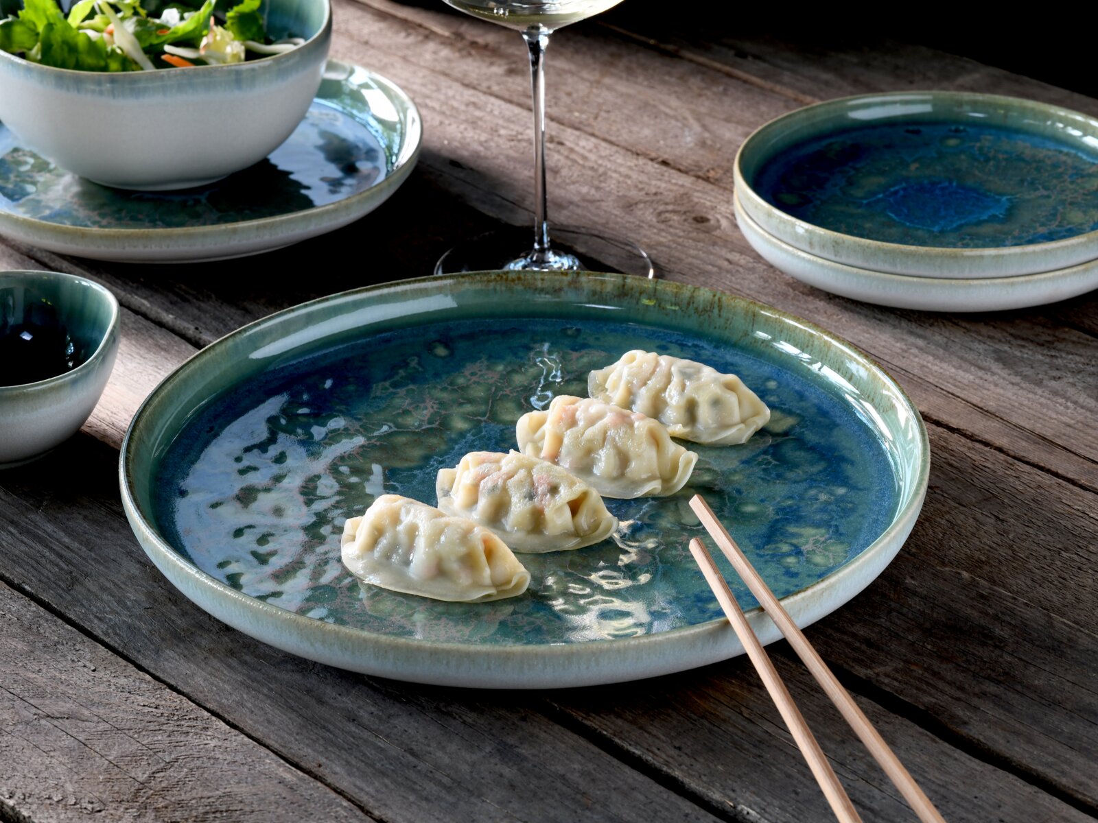 Blau-grüner, glasierter Keramikteller mit vier Teigtaschen und Holzstäbchen auf rustikalem Holztisch, aufgenommen in leicht schräger Draufsicht; im Hintergrund Schale mit Salat, kleines Schälchen und Weinglas.