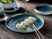 Blau-grüner, glasierter Keramikteller mit vier Teigtaschen und Holzstäbchen auf rustikalem Holztisch, aufgenommen in leicht schräger Draufsicht; im Hintergrund Schale mit Salat, kleines Schälchen und Weinglas.