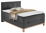 Boxspringbett CATANIA in dunklem Stoff mit Holzfüßen, seitliche Perspektive, dekoriert mit Kissen und Decke.