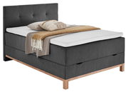 Boxspringbett CATANIA in dunklem Stoff mit Holzfüßen, seitliche Perspektive, dekoriert mit Kissen und Decke.
