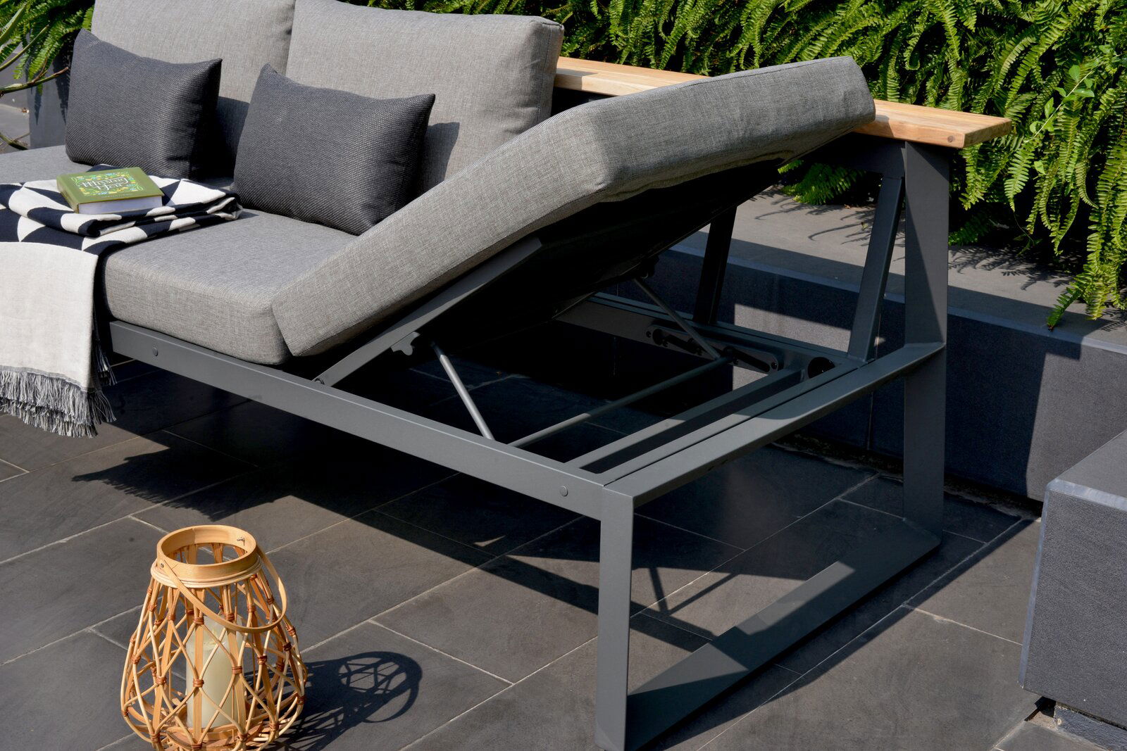Graue Sitzbank mit verstellbarer Rückenlehne, seitliche Perspektive, dekoriert mit Kissen, Decke und Buch, umgeben von Pflanzen.
