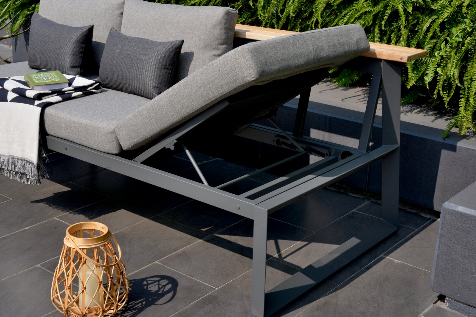 Graue Sitzbank mit verstellbarer Rückenlehne, seitliche Perspektive, dekoriert mit Kissen, Decke und Buch, umgeben von Pflanzen.