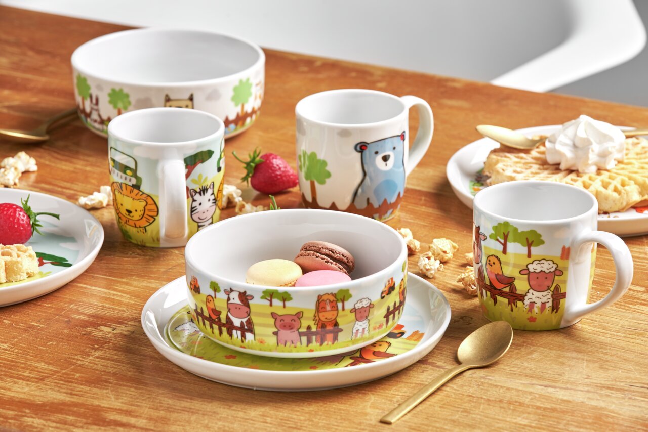 Van Well Kinderset 3-tlg. BAUERNHOF Kinderset mit Bauernhof-Motiven, bestehend aus Tassen, Schalen und Tellern, auf einem Holztisch arrangiert. Perspektive von oben.