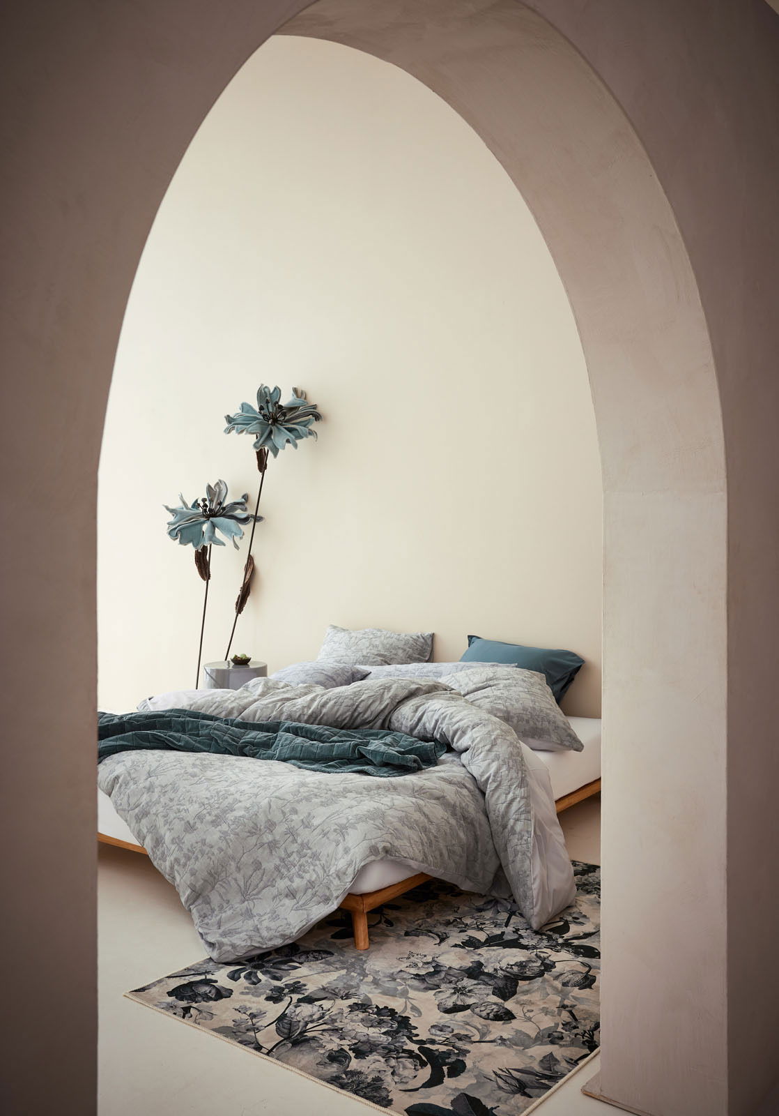 Blick durch einen Bogen auf ein stilvoll eingerichtetes Schlafzimmer mit Bettwäsche in sanften Grautönen und floralen Mustern, dekoriert mit großen blauen Blumen an der Wand.