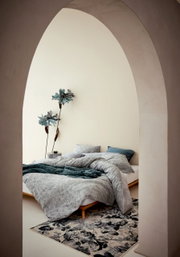 Blick durch einen Bogen auf ein stilvoll eingerichtetes Schlafzimmer mit Bettwäsche in sanften Grautönen und floralen Mustern, dekoriert mit großen blauen Blumen an der Wand.