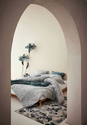 Blick durch einen Bogen auf ein stilvoll eingerichtetes Schlafzimmer mit Bettwäsche in sanften Grautönen und floralen Mustern, dekoriert mit großen blauen Blumen an der Wand.