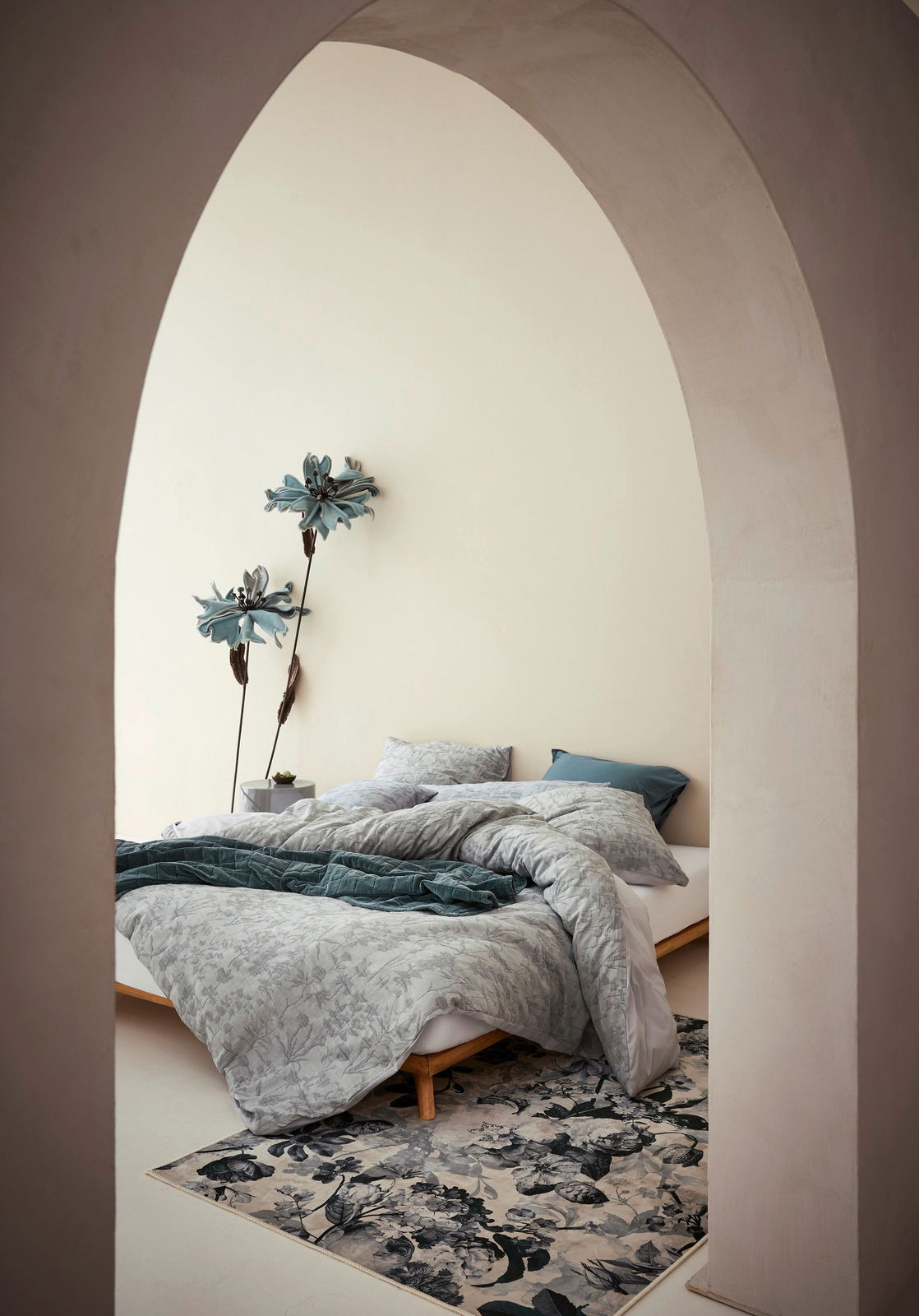 Blick durch einen Bogen auf ein stilvoll eingerichtetes Schlafzimmer mit Bettwäsche in sanften Grautönen und floralen Mustern, dekoriert mit großen blauen Blumen an der Wand.