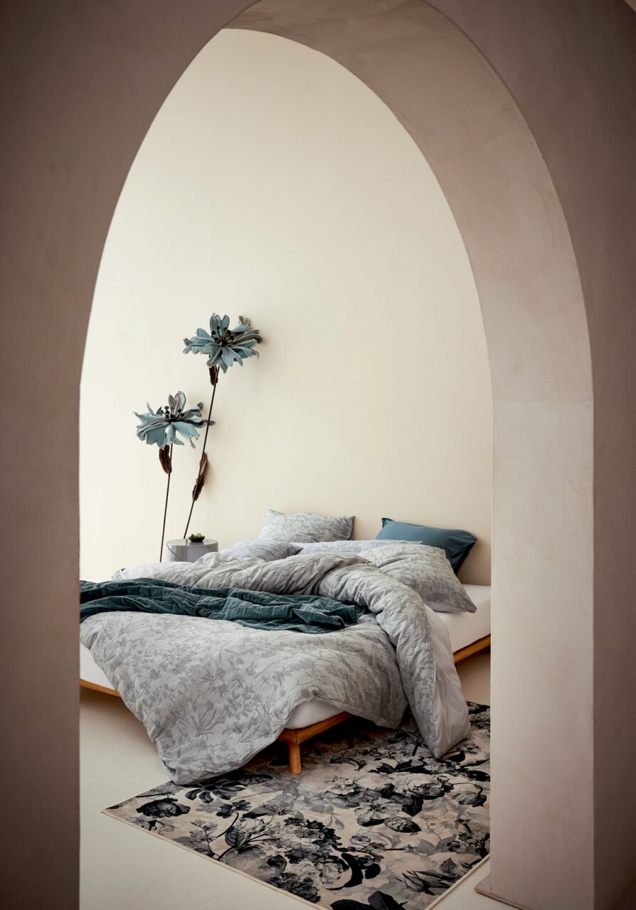 Essenza Home Beddengoed MARIBEL Uitzicht door een boog op een stijlvol ingerichte slaapkamer met beddengoed in zachte grijstinten en bloemmotieven, versierd met grote blauwe bloemen aan de muur.