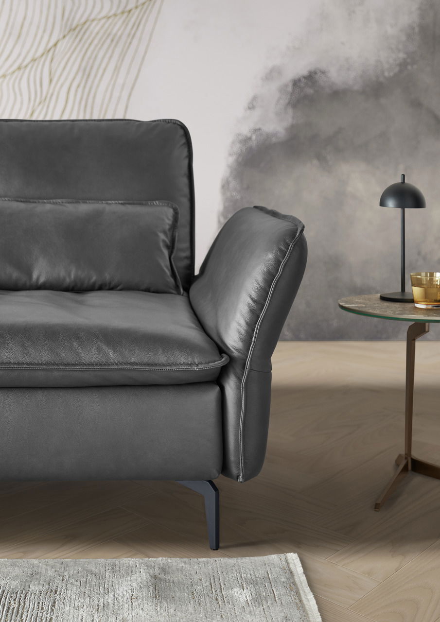 Detailansicht eines modernen, dunkelgrauen Longchairs mit eleganten Metallfüßen, im Vordergrund ein Beistelltisch mit Lampe und Glas. Perspektive von der Seite.