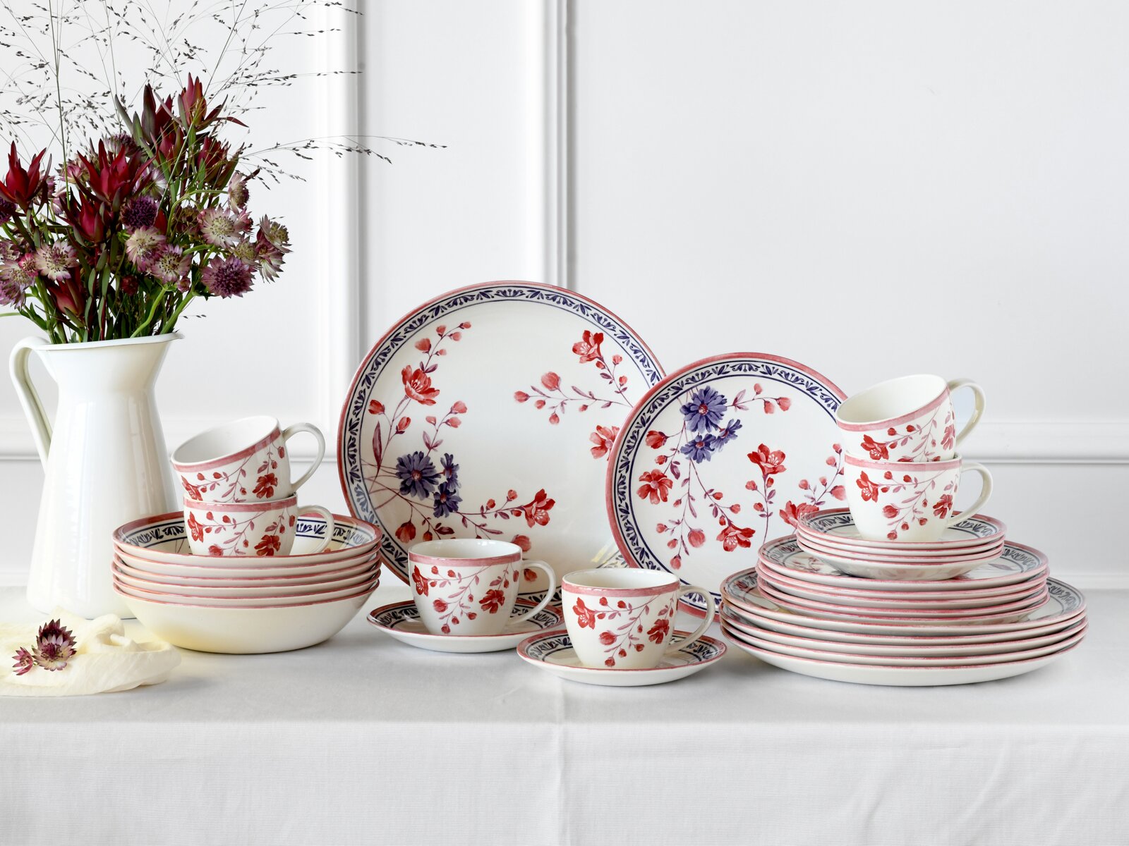 creaTable Kombiservice 30-tlg COTTAGE Porzellan-Geschirrset mit roten und blauen Blumenmotiven, Teller, Schalen und Tassen auf weißer Tischdecke arrangiert, frontal in leicht schräger Perspektive fotografiert; links eine weiße Vase mit Blumen