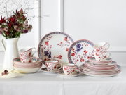 Porzellan-Geschirrset mit roten und blauen Blumenmotiven, Teller, Schalen und Tassen auf weißer Tischdecke arrangiert, frontal in leicht schräger Perspektive fotografiert; links eine weiße Vase mit Blumen
