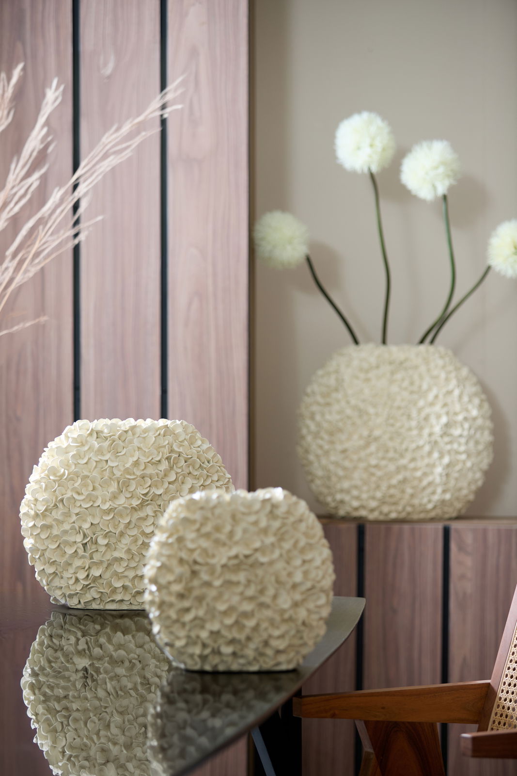 Zwei cremefarbene Vasen mit blütenartiger Struktur auf einem Tisch, im Hintergrund eine größere Vase mit dekorativen Blumen, seitliche Perspektive.