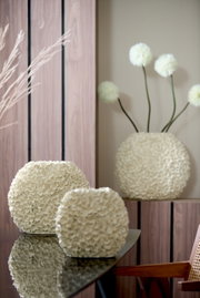 Zwei cremefarbene Vasen mit blütenartiger Struktur auf einem Tisch, im Hintergrund eine größere Vase mit dekorativen Blumen, seitliche Perspektive.