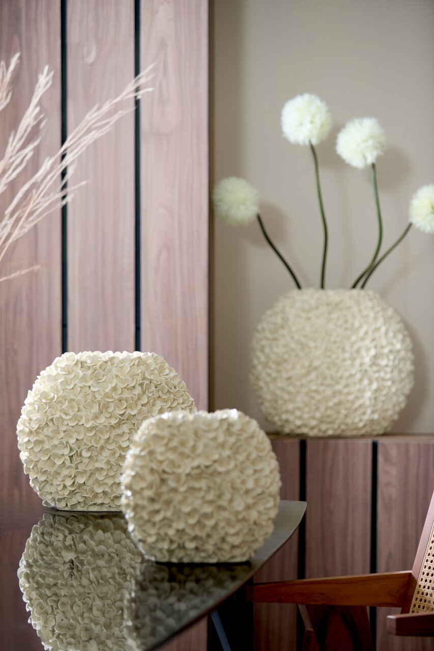 Zwei cremefarbene Vasen mit blütenartiger Struktur auf einem Tisch, im Hintergrund eine größere Vase mit dekorativen Blumen, seitliche Perspektive.