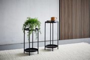 Zwei schwarze Blumensäulen aus Metall, eine mit einer Pflanze und die andere mit einer Tasse und Büchern, auf einem hellen Teppich vor einer Wand mit Holzvertäfelung, seitliche Perspektive.