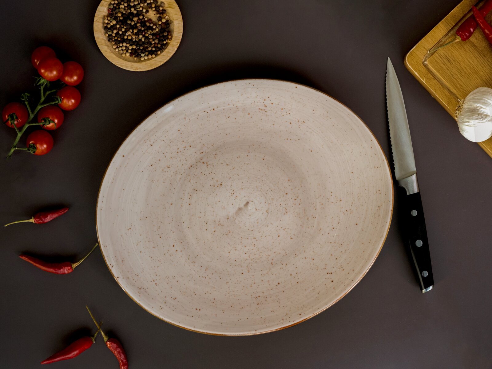 Großer, flacher, gesprenkelter Keramikteller in Beige auf dunklem Untergrund, umgeben von Tomaten, Chili, Pfefferkörnern, Knoblauch und Messer; Draufsicht von oben
