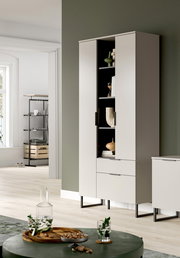 Moderner hoher Wohnzimmerschrank in Hellgrau mit schwarzen Metallfüßen, zwei Türen und Schublade, eine Tür geöffnet mit sichtbaren Regalfächern und Deko, in heller Wohnraum-Szene; Perspektive: leicht schräg von vorn/rechts.