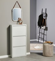 Modernes, weißes Schuhschrank-/Klappschrankmöbel mit drei Klappen im Flur, darüber ein abgerundeter Wandspiegel mit Lederaufhängung; seitliche Frontansicht in einer eingerichteten Ecke mit Leitergarderobe und Pouf.