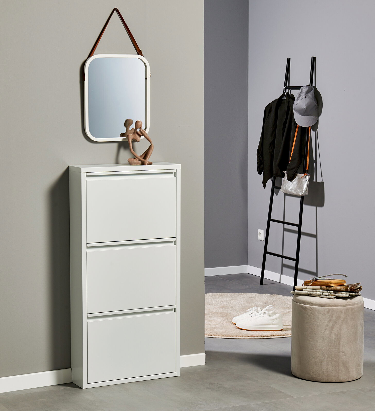 Modernes, weißes Schuhschrank-/Klappschrankmöbel mit drei Klappen im Flur, darüber ein abgerundeter Wandspiegel mit Lederaufhängung; seitliche Frontansicht in einer eingerichteten Ecke mit Leitergarderobe und Pouf.