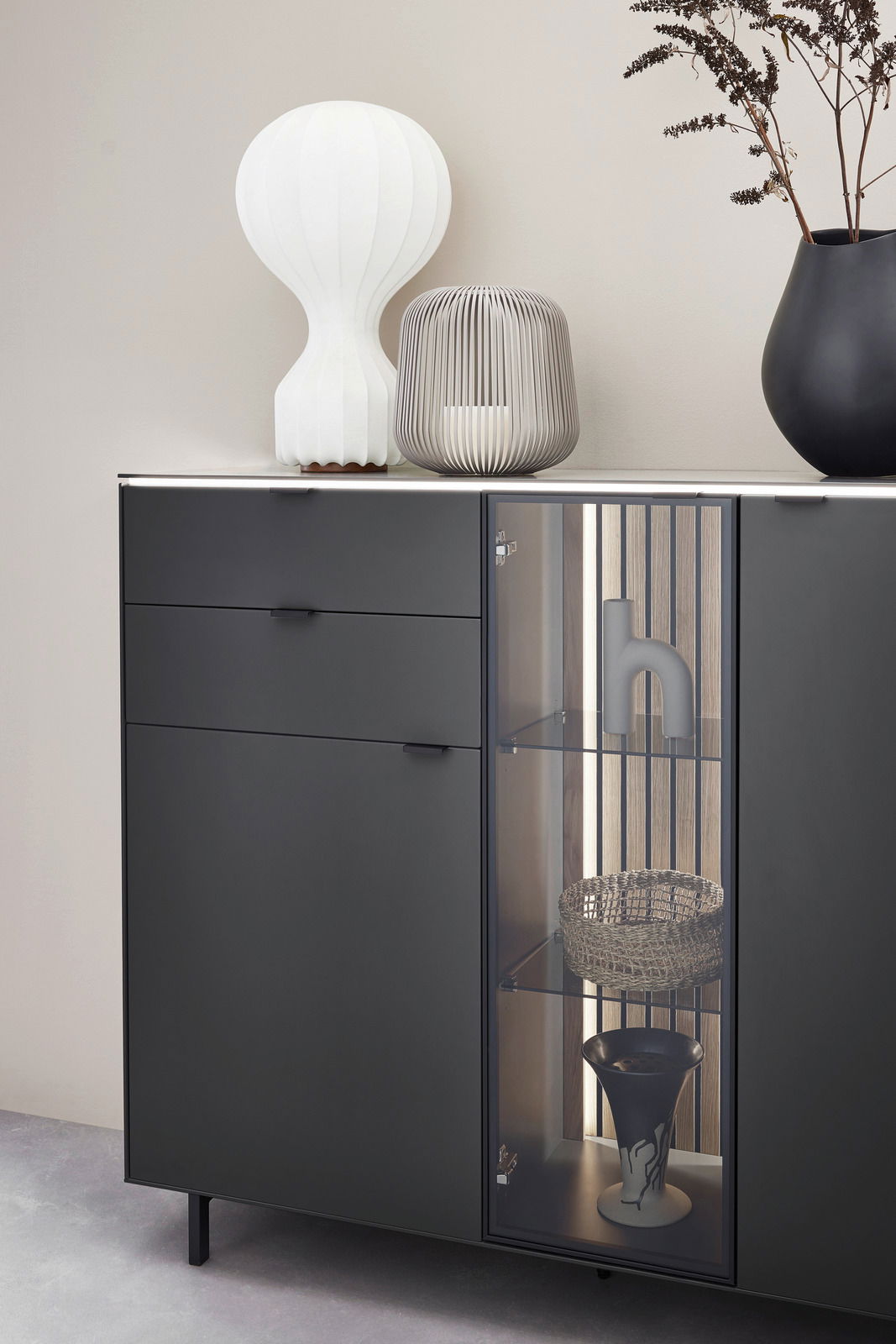 Modernes Highboard in dunklem Design mit Glasvitrine, dekoriert mit einer weißen Lampe, einer grauen Laterne und einer schwarzen Vase. Frontale Perspektive.