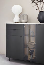 Modernes Highboard in dunklem Design mit Glasvitrine, dekoriert mit einer weißen Lampe, einer grauen Laterne und einer schwarzen Vase. Frontale Perspektive.
