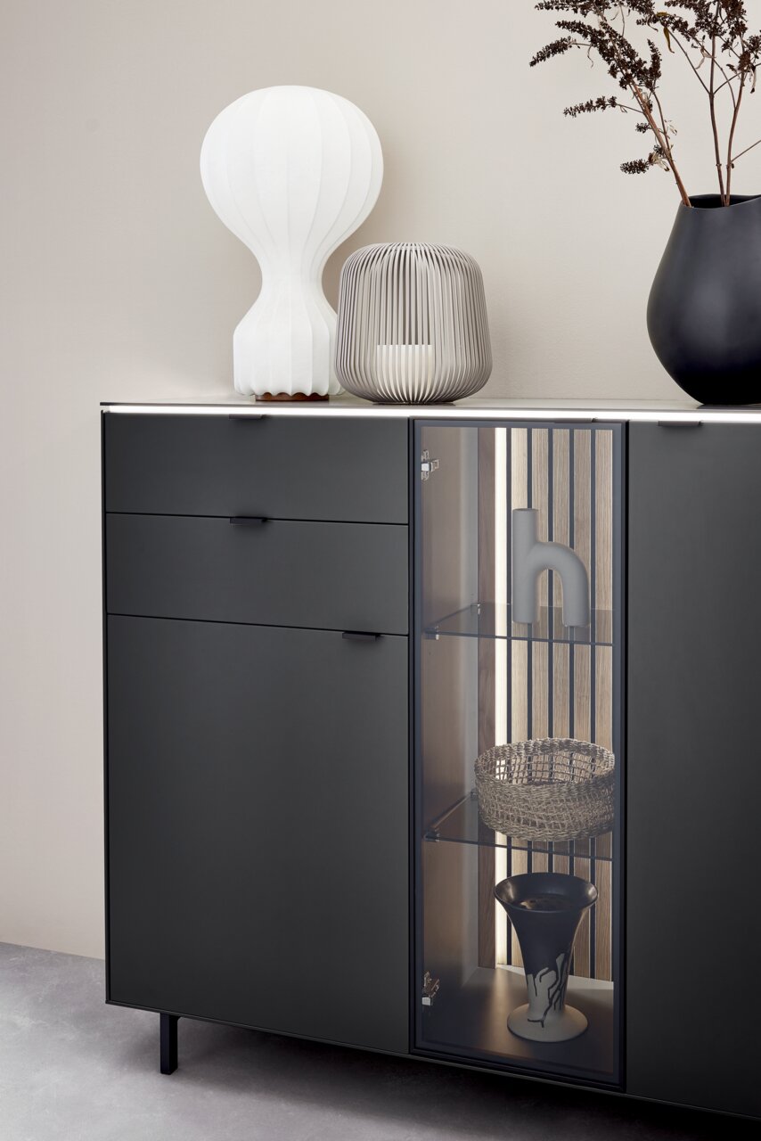 Modernes Highboard in dunklem Design mit Glasvitrine, dekoriert mit einer weißen Lampe, einer grauen Laterne und einer schwarzen Vase. Frontale Perspektive.